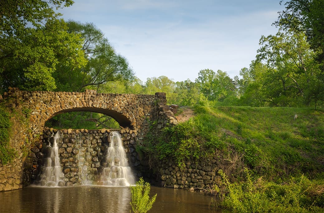Stone arch bridge with waterfall in a park, ideal for families in Winston-Salem.