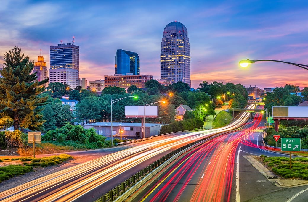 Winston-Salem skyline at dusk with highway traffic, comparing living costs and lifestyle.