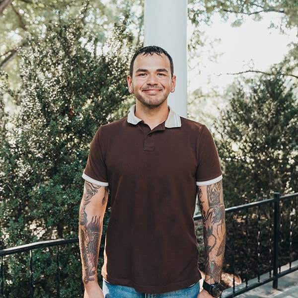 Dylan McDonald Dylan McDonald, a man with tattoos, smiles outdoors in front of greenery.